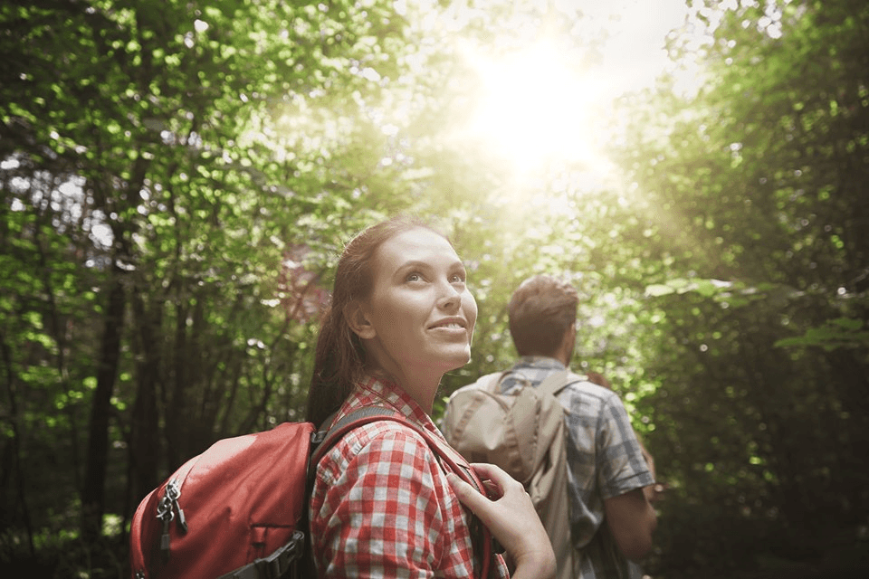 A woman with a backpack smiles while walking through a sunlit forest with a man behind her.
