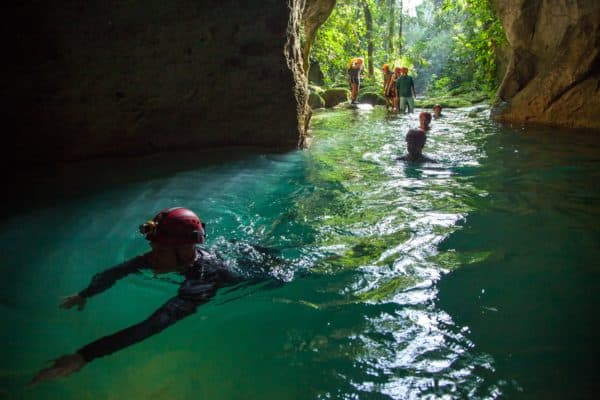 A group of people explores a turquoise water cave while one person swims in the foreground.