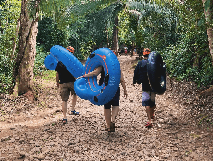 Three people walking on a rocky path, each carrying an inner tube amidst lush greenery.
