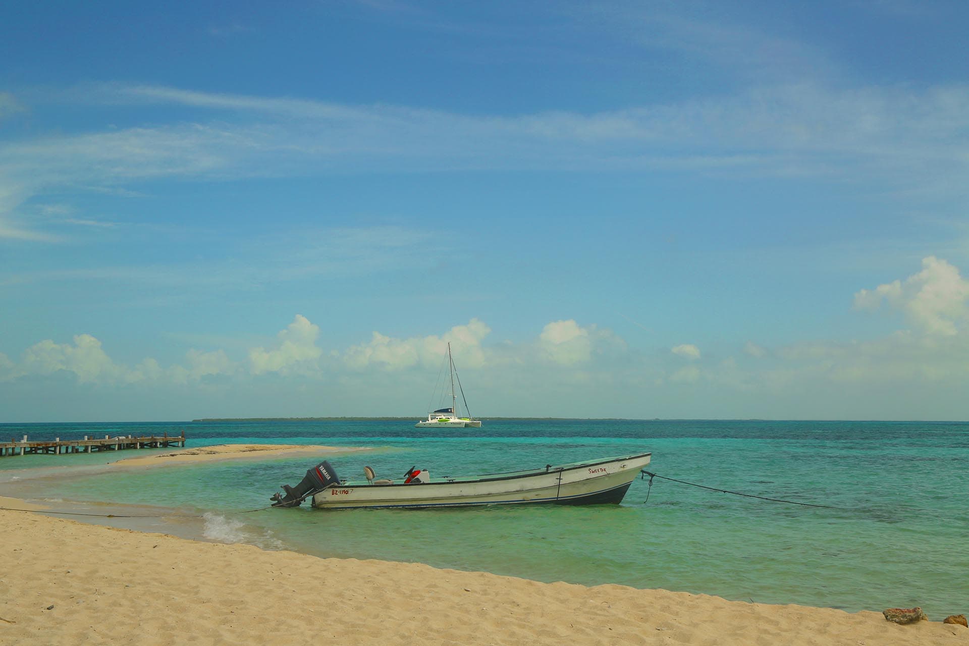 A small boat is anchored on a sandy beach with calm turquoise waters and a distant sailboat under a clear blue sky.