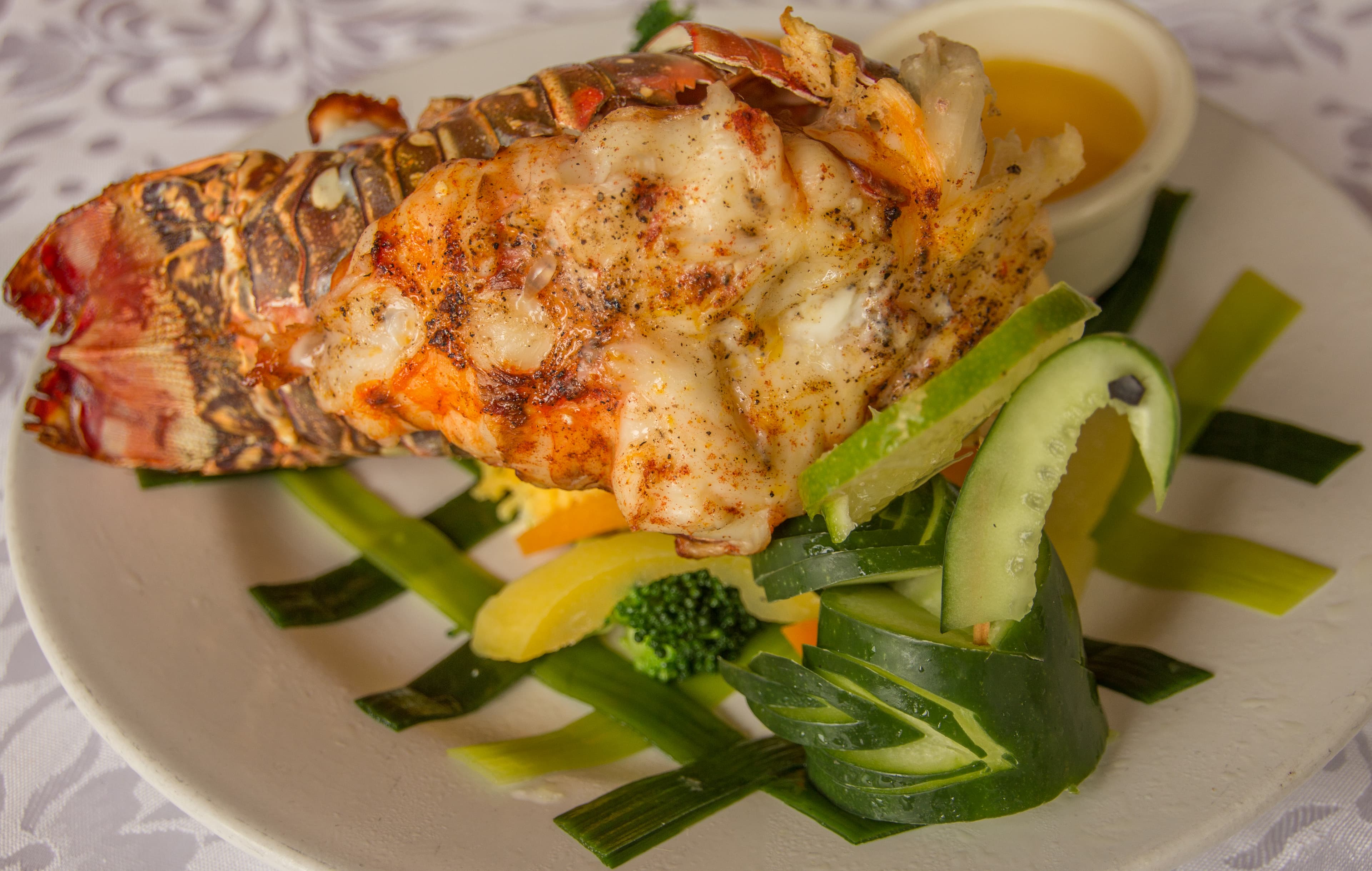 Grilled lobster tail served on a bed of colorful vegetables with a side of dipping sauce.