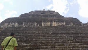 A person stands at the base of a steep, ancient pyramid under a cloudy sky.