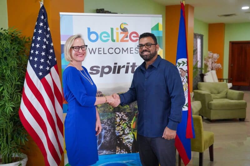 A man and woman shake hands in front of a "Belize Welcomes Spirit" banner, with flags of Belize and the U.S. in the background.