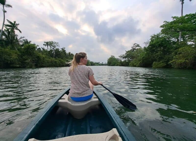 A woman paddles a kayak on a calm river surrounded by lush trees and clouds.