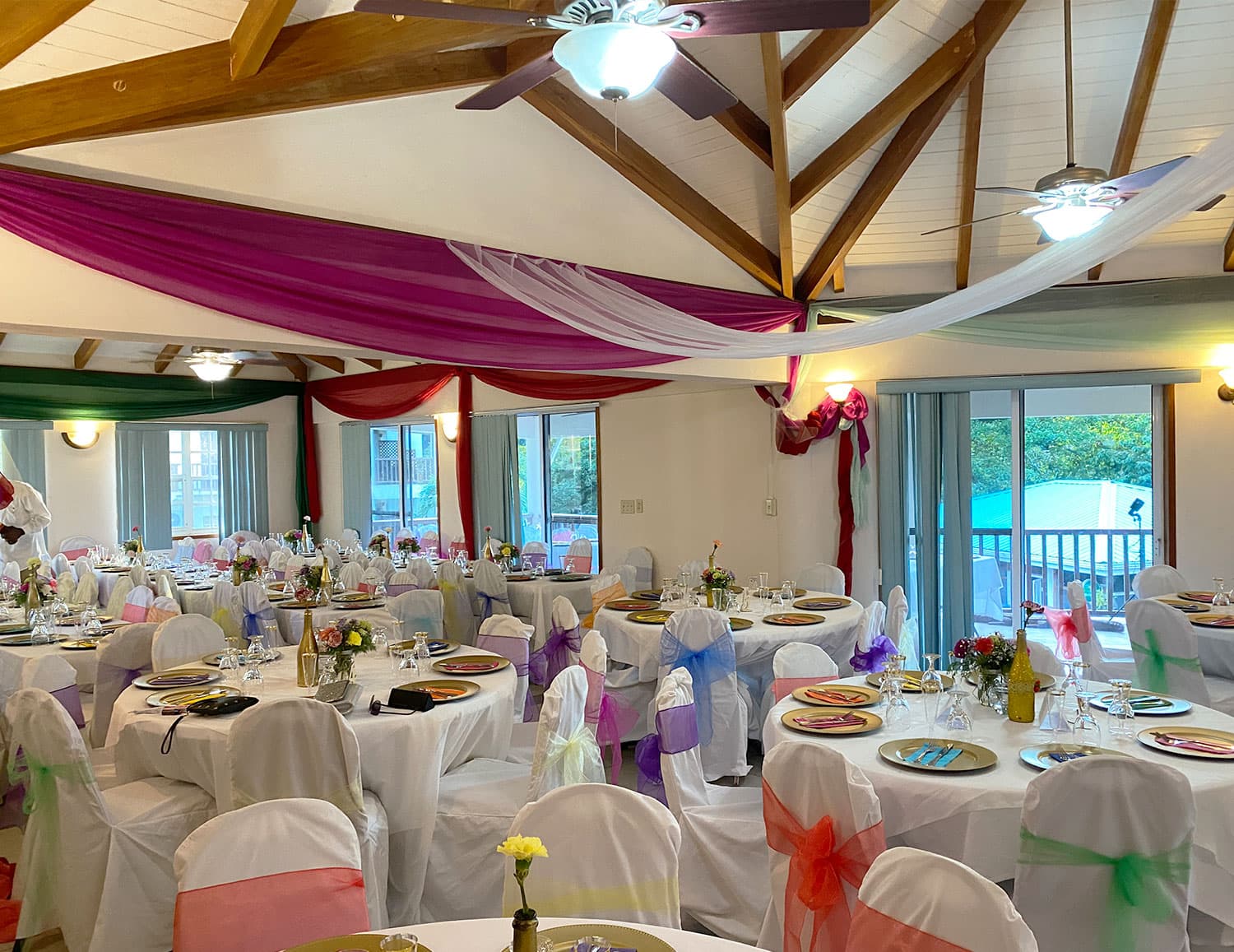A beautifully decorated dining area featuring tables set with colorful chair sashes and floral centerpieces under an elegant wooden ceiling.