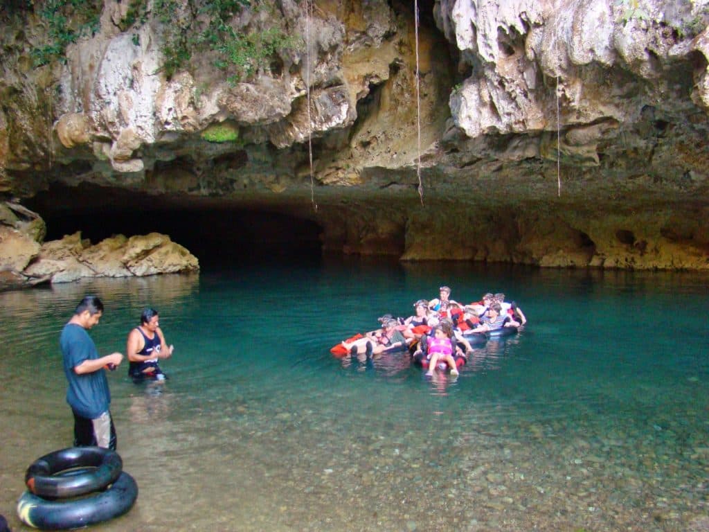 A group of people floats in a clear water cave while one person stands on the shore.
