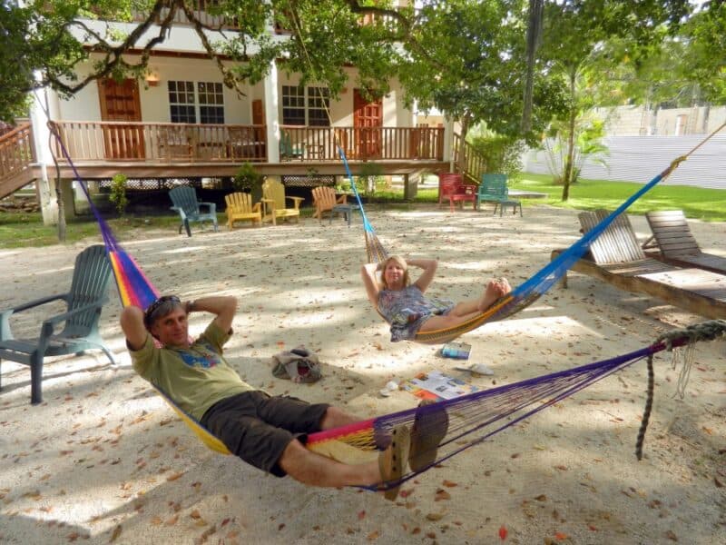 Two people relax in colorful hammocks on a sandy ground beneath trees, with a house and chairs in the background.