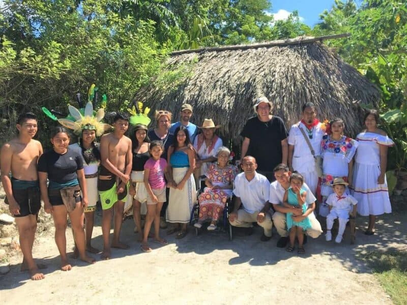 A diverse group of people, dressed in traditional attire, poses in front of a thatched-roof hut in a lush green setting.