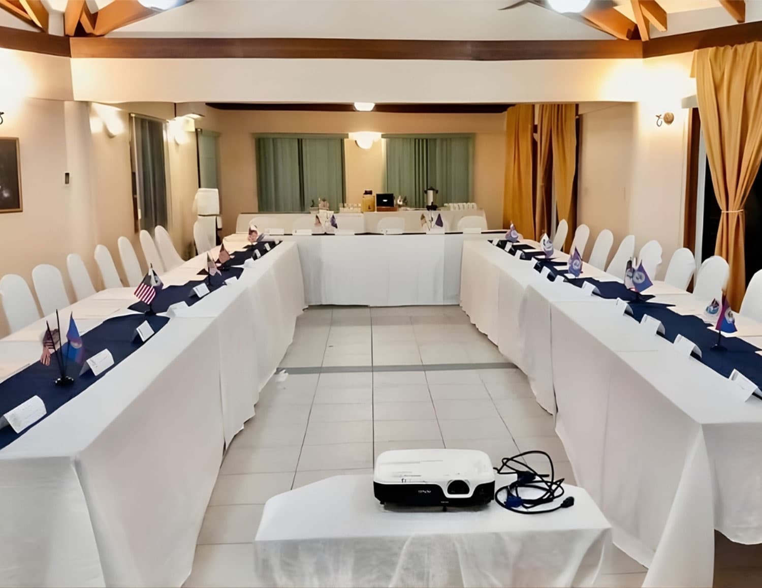 A conference room set up with two long tables, each decorated with flags and nameplates, and a projector in the center.