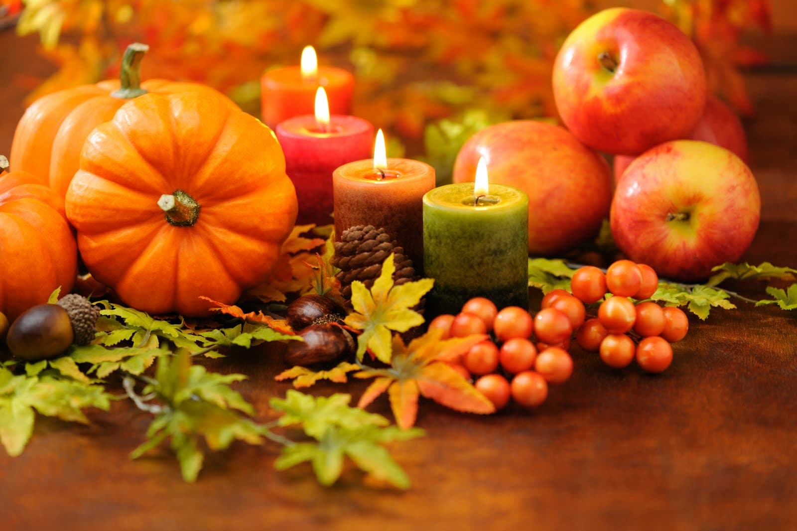 A festive arrangement of pumpkins, apples, colorful candles, and autumn leaves on a wooden surface.