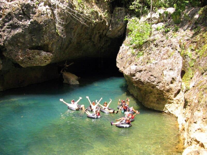 Group of people in inner tubes enjoying a sunny day in a natural pool surrounded by rocky cliffs and lush greenery.