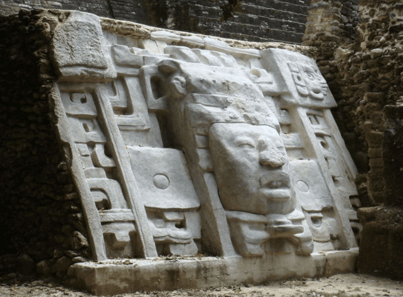 A carved stone relief with a face and intricate designs, set against a mossy, historical backdrop.