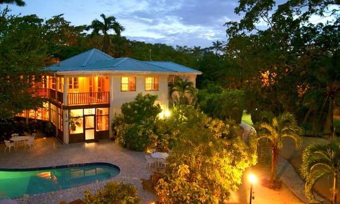 A two-story house surrounded by lush greenery and a swimming pool, illuminated at dusk.