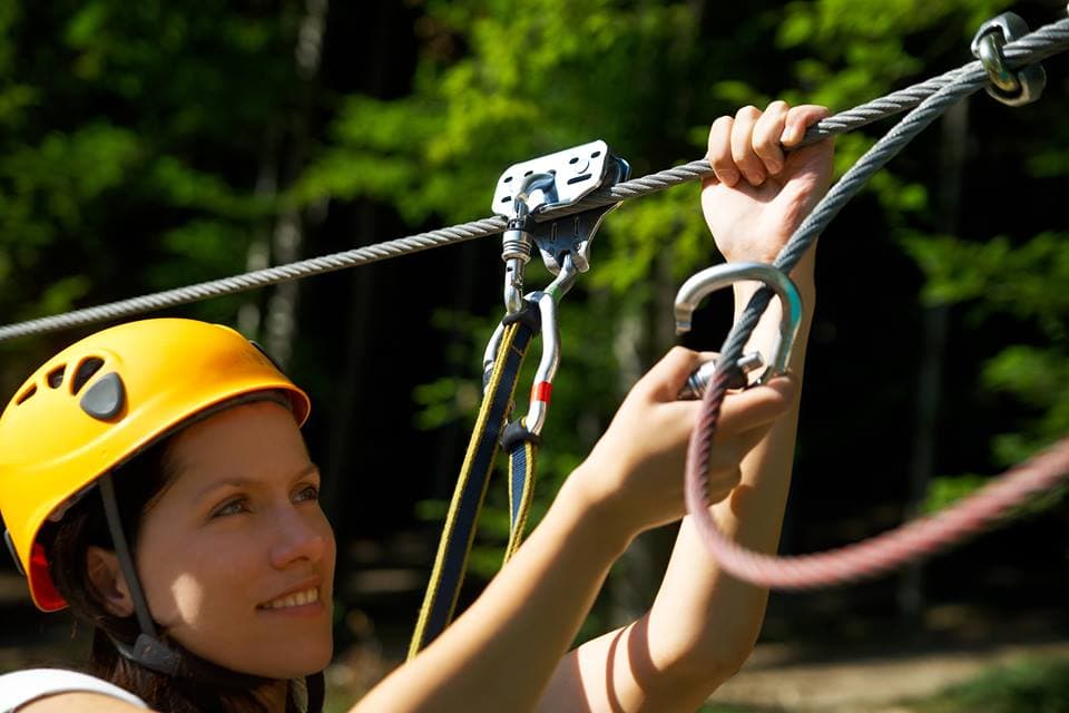 A woman in a helmet adjusts a safety clip on a zip line.