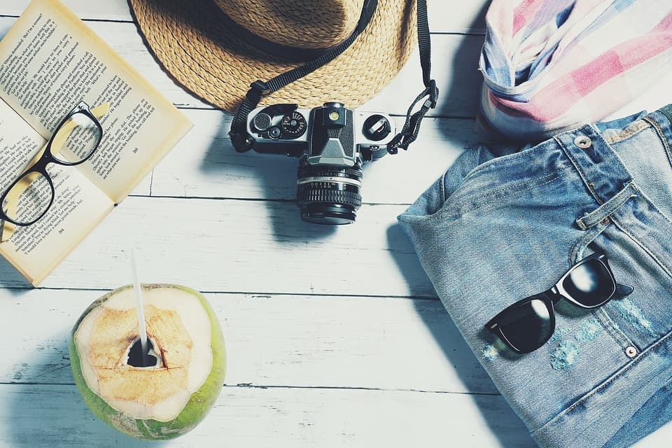 Flat lay of a straw hat, camera, sunglasses, book, denim shorts, and a coconut drink on a wooden surface.