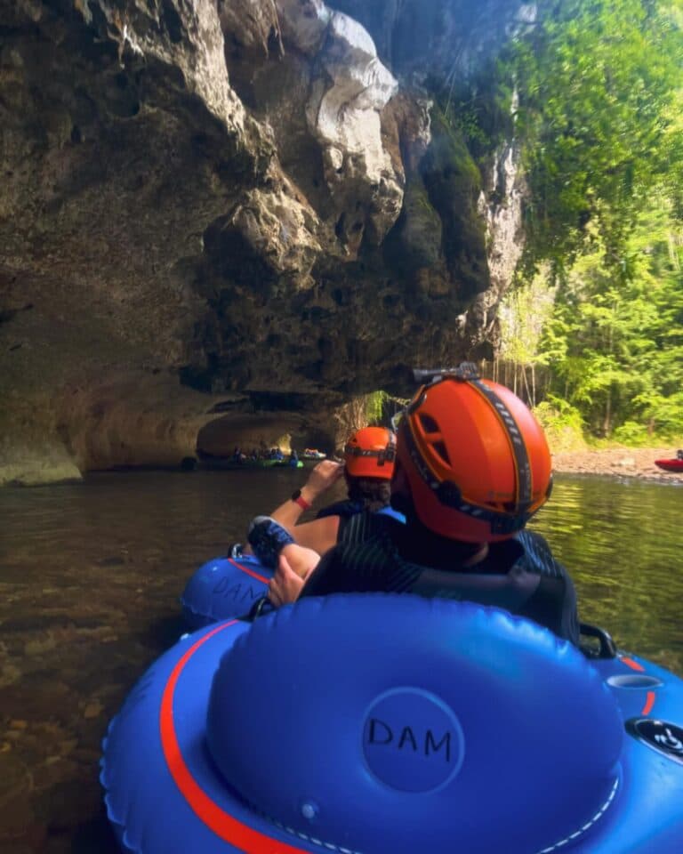Two people in orange helmets float on inner tubes through a rocky cave surrounded by lush greenery.