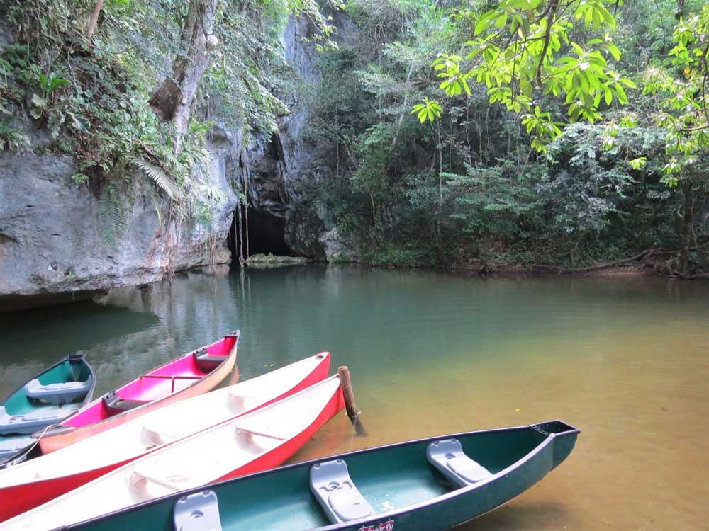 A group of colorful canoes docked by a tranquil river surrounded by lush greenery and rocky cliffs.