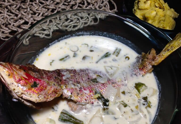 A bowl of creamy fish soup featuring a whole red snapper and assorted vegetables, accompanied by a side of mashed plantains.