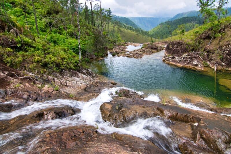 A serene landscape featuring a flowing stream and tranquil pool surrounded by lush greenery and hills.