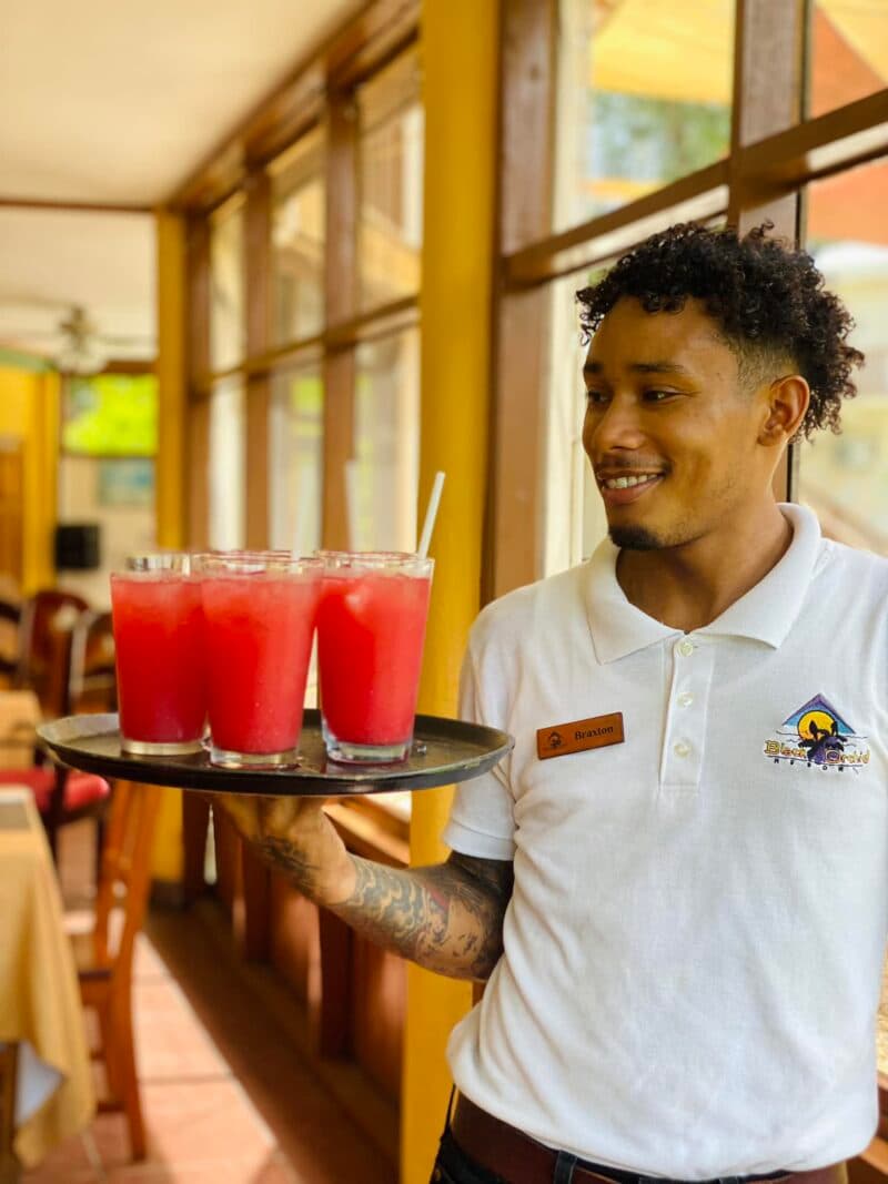 A smiling waiter holds a tray of bright red drinks in a sunny restaurant interior.