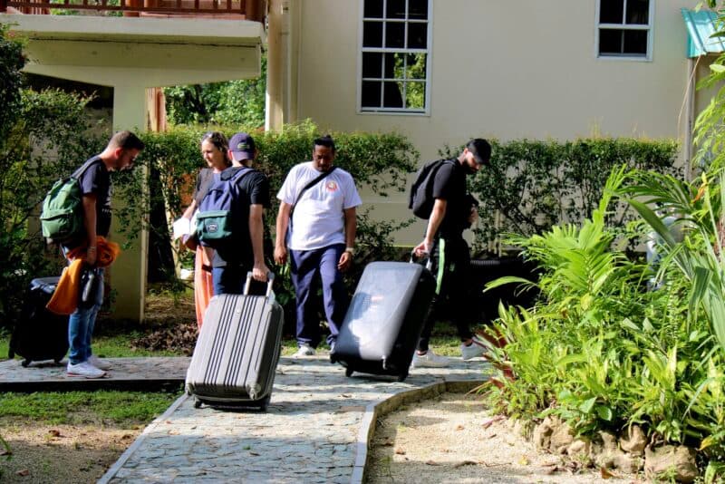 A group of four people is seen walking with suitcases along a path near a building surrounded by greenery.