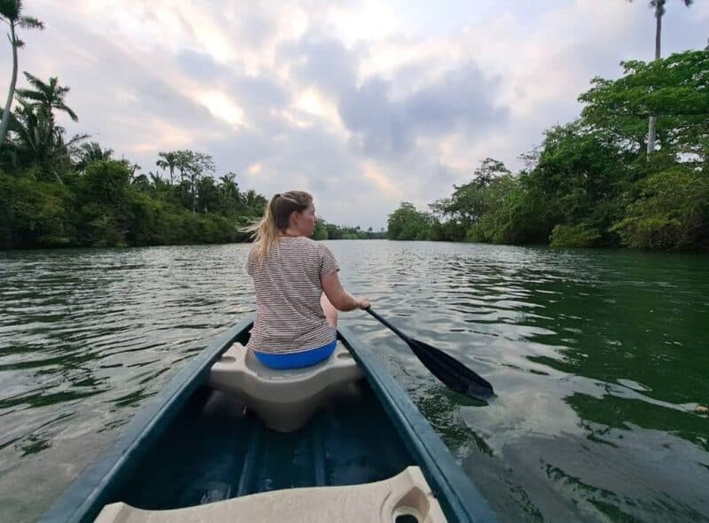 A person paddles in a canoe on a calm river surrounded by lush greenery and a cloudy sky.