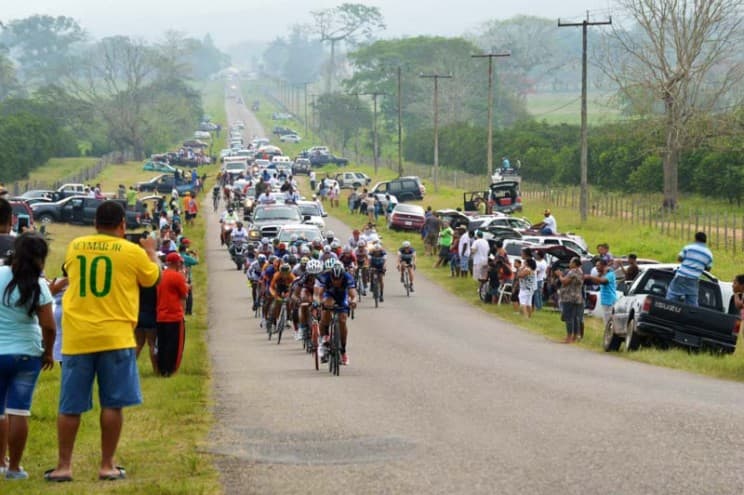 A group of cyclists races down a rural road while a crowd and parked cars line the sides.