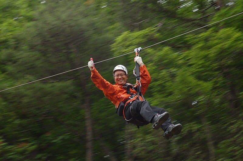 A person in an orange jacket enjoys zip-lining through a forested area, giving a thumbs up.