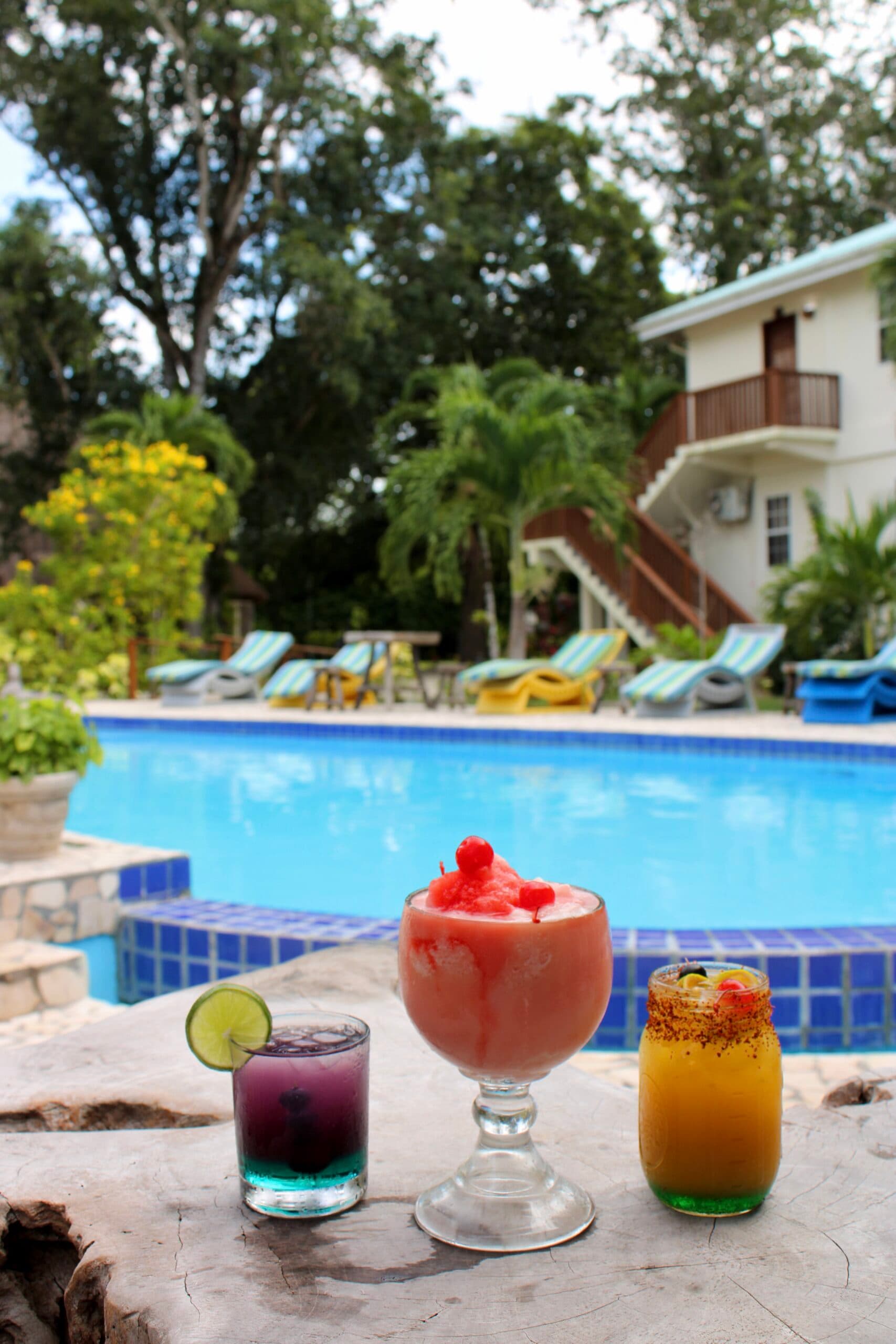 Three colorful cocktails sit on a table by a swimming pool surrounded by lush greenery.