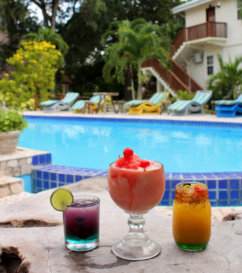 Three colorful cocktails are displayed on a table by a swimming pool surrounded by tropical plants and lounge chairs.
