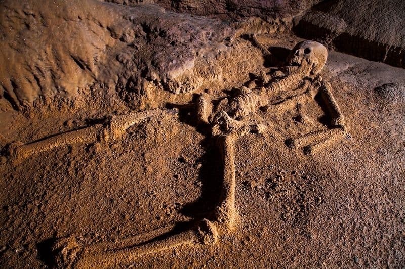 A human skeleton partially buried in sandy sediment inside a cave.