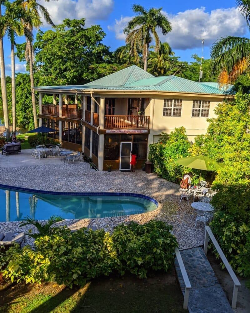 A two-story house with a teal roof surrounded by palm trees, a swimming pool, and outdoor seating areas.
