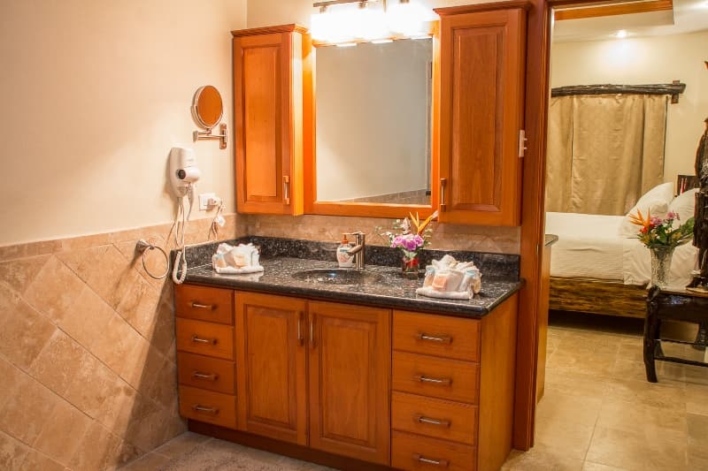 A modern bathroom featuring a dark countertop, wooden cabinetry, and a mirror, with a glimpse of a bedroom in the background.