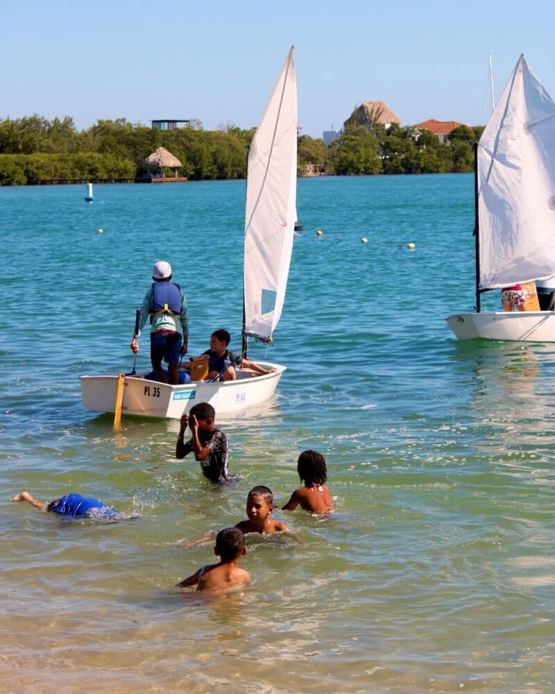 Children swim and play in shallow water while others sail on small boats nearby.