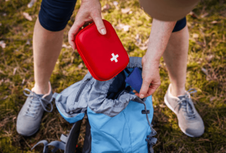 A person adds a red first aid kit to a blue backpack while outdoors.
