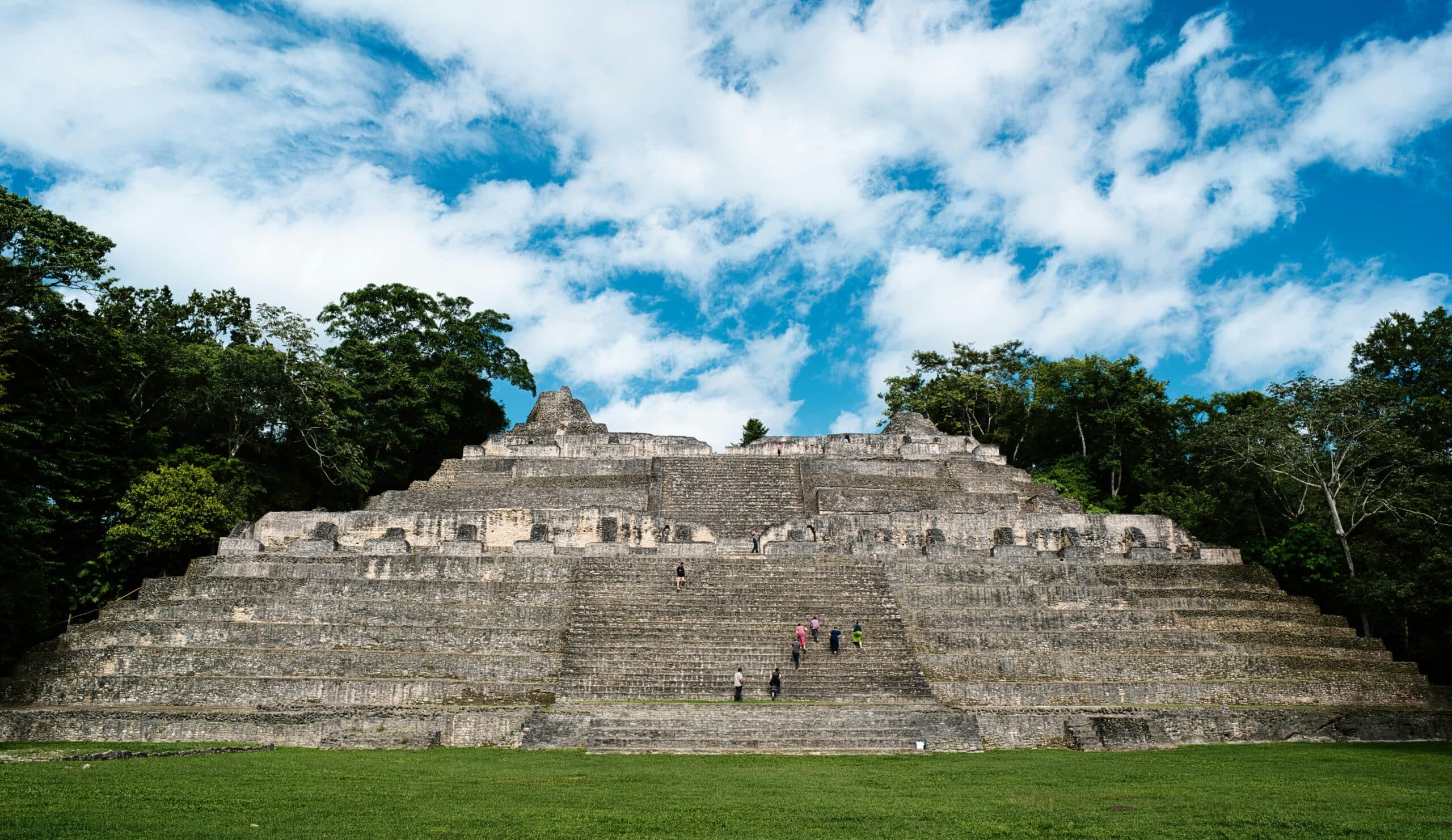 A large, ancient Mayan pyramid surrounded by lush greenery under a blue sky with scattered clouds.
