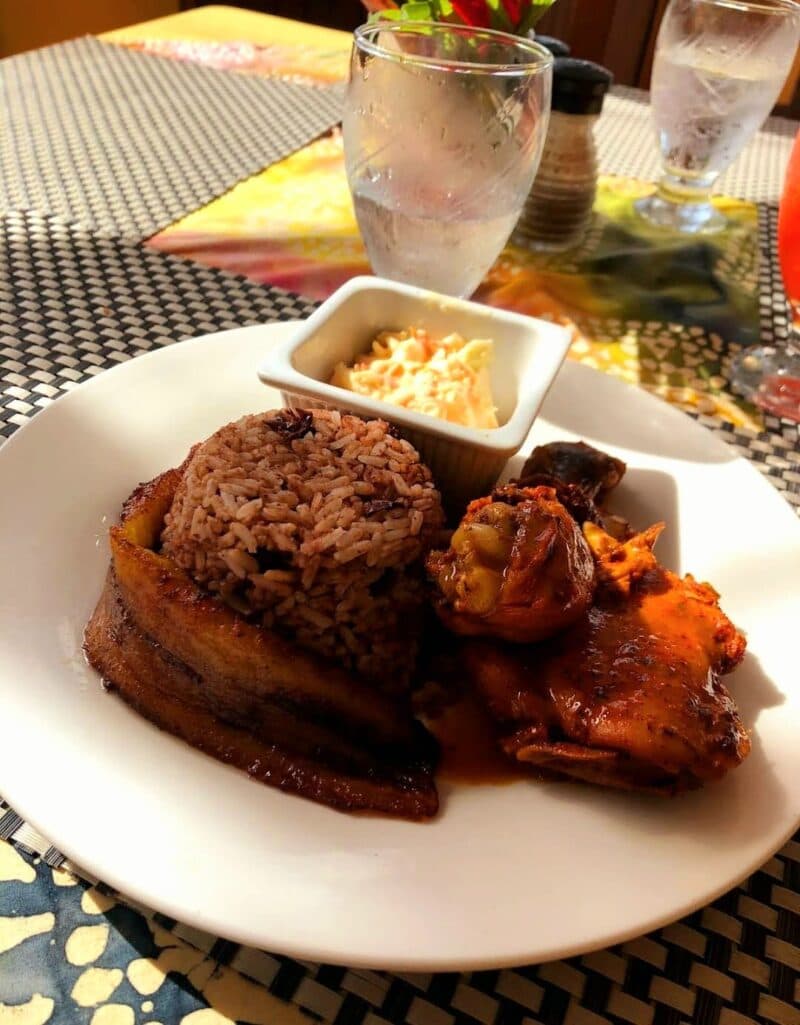 A plate of rice, fried plantains, chicken, and a small serving of coleslaw, with drinks in the background.