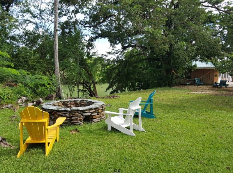 A cozy outdoor scene featuring colorful chairs around a stone fire pit, set in a lush green yard near a body of water.