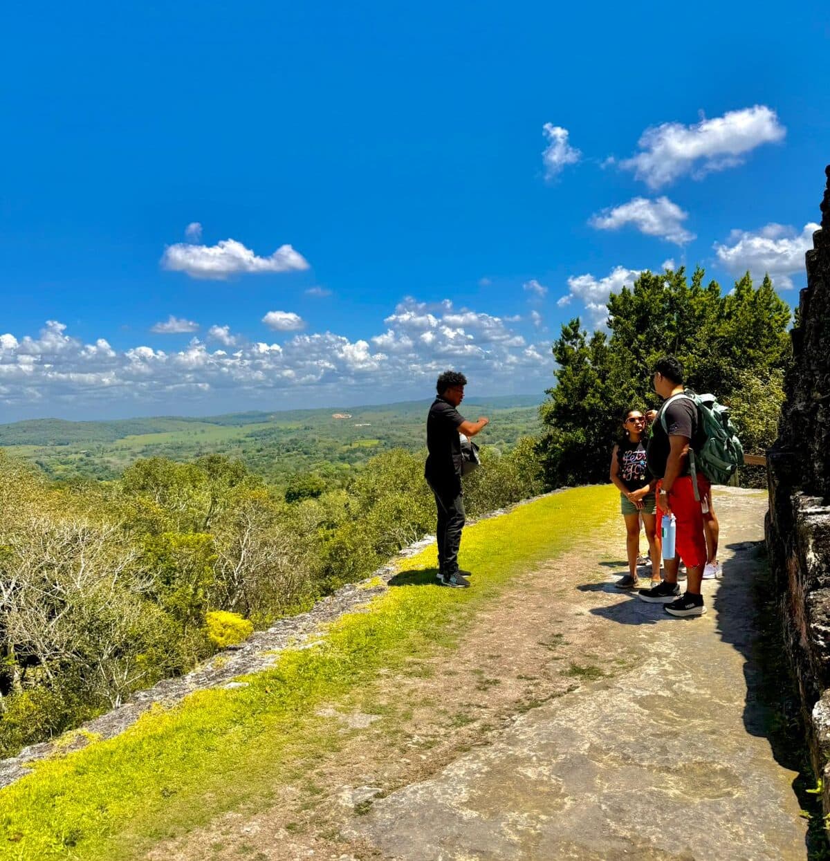 Two people converse with a tour guide on a sunny hilltop overlooking a lush valley.
