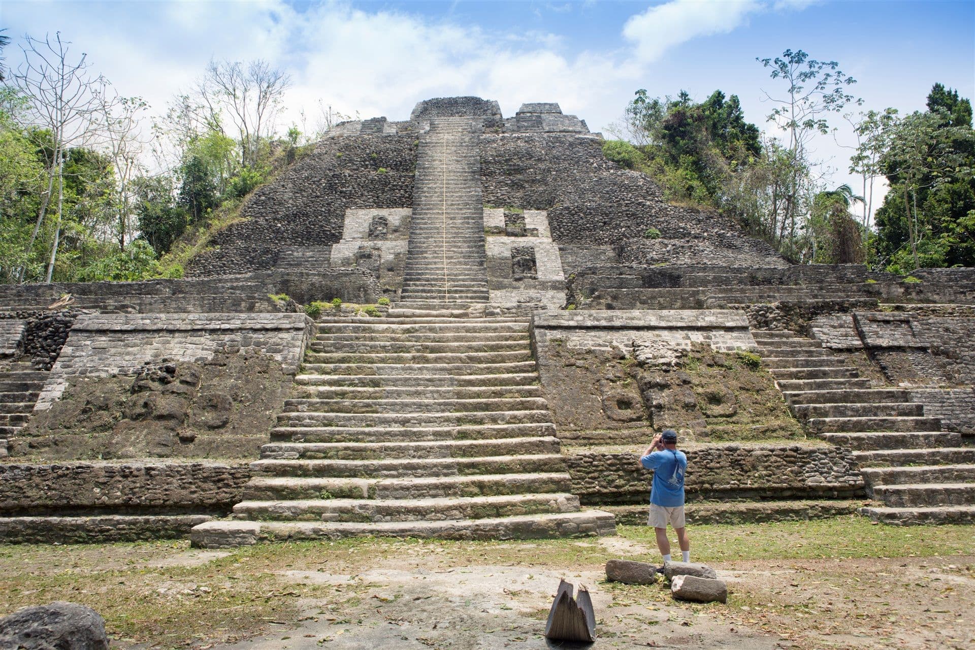 A person stands in front of a large ancient pyramid, taking a photo while surrounded by lush greenery.