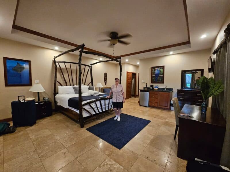 A woman stands in a spacious hotel room featuring a four-poster bed, a kitchenette, and a desk.