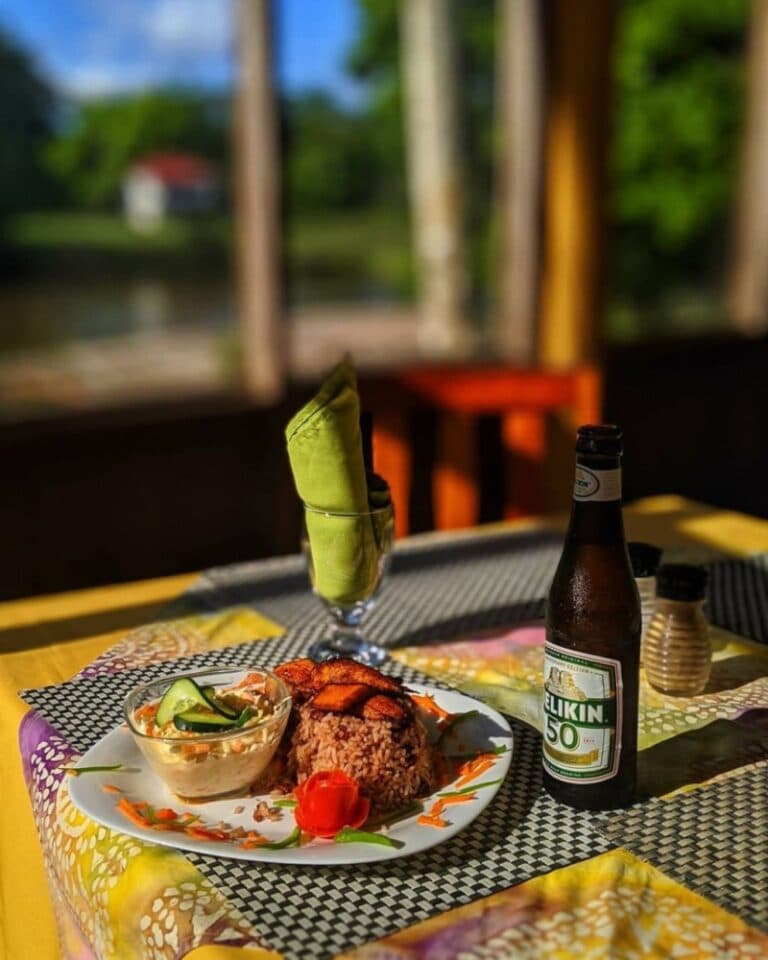 A colorful plate of rice and vegetables alongside a beer, set on a table with vibrant linens and a view of greenery.