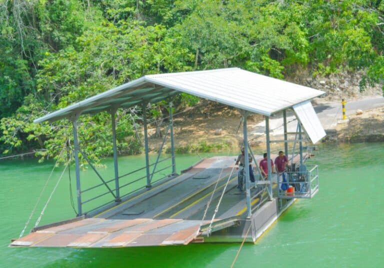 A metal ferry with passengers is docked on a green river surrounded by lush trees.
