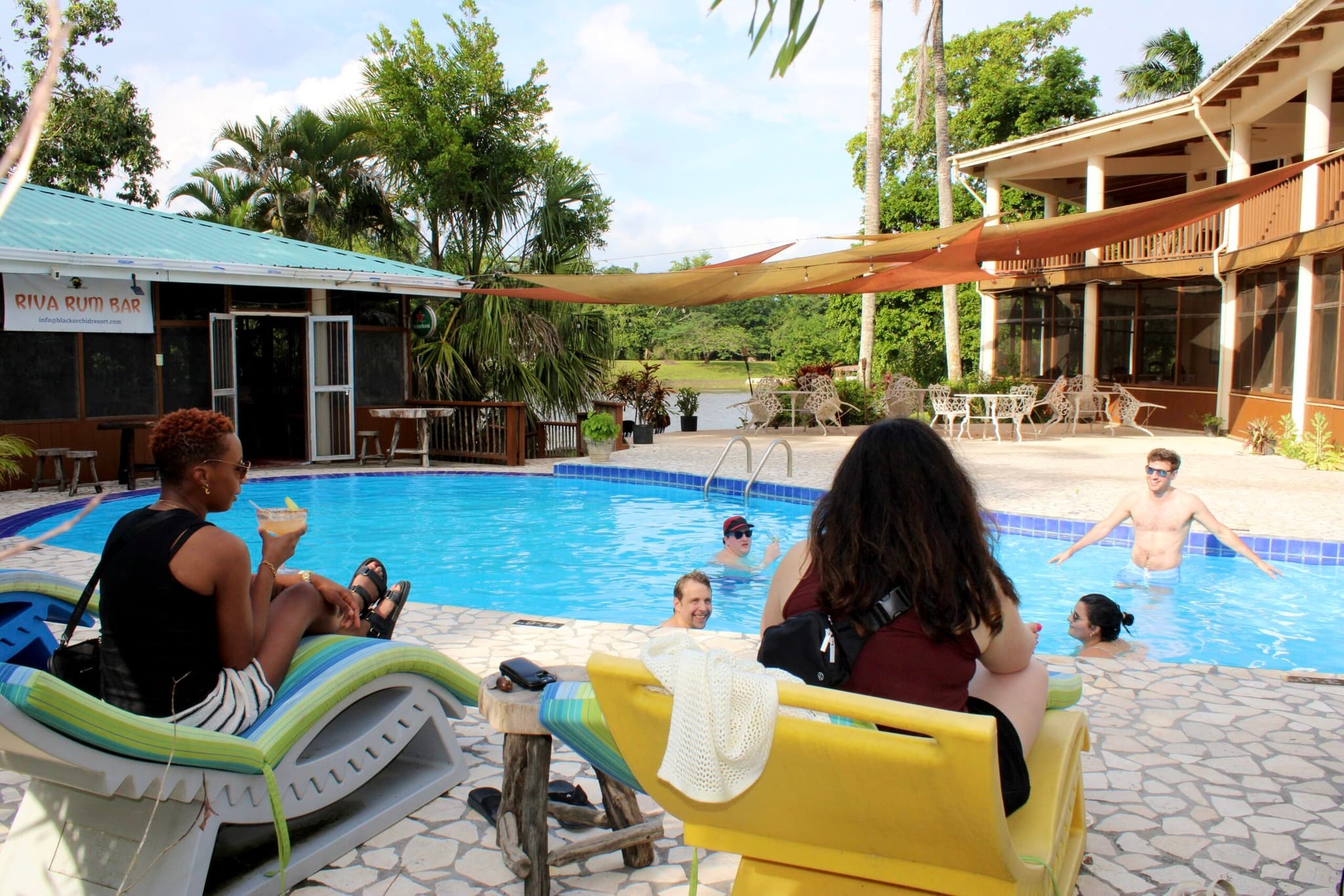 People relax by a pool surrounded by lush greenery and a casual outdoor setting.