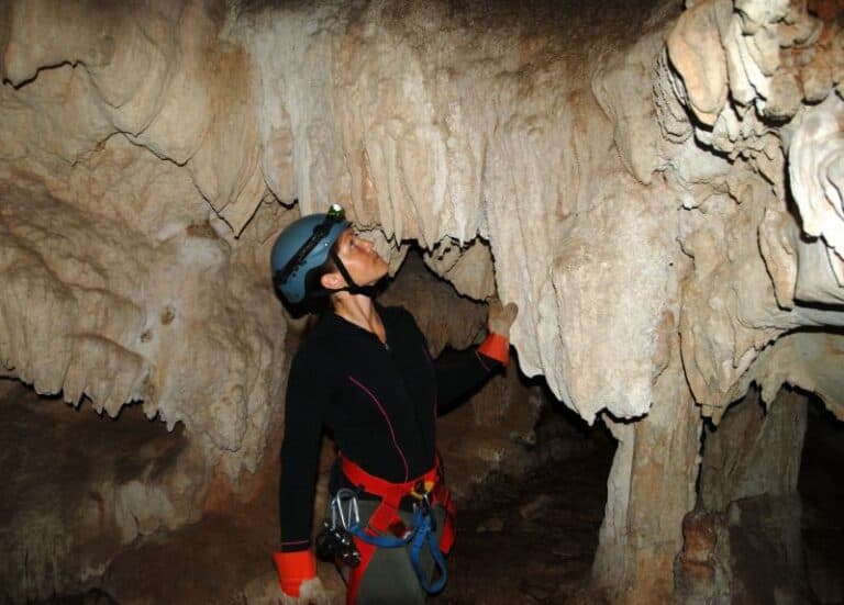 A person wearing a helmet explores a cave, gazing up at the stalactites above.