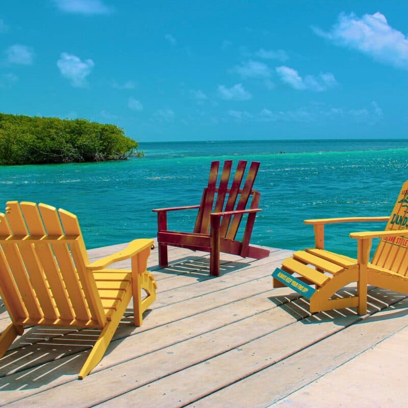 Three colorful Adirondack chairs face a tranquil turquoise sea under a bright blue sky.