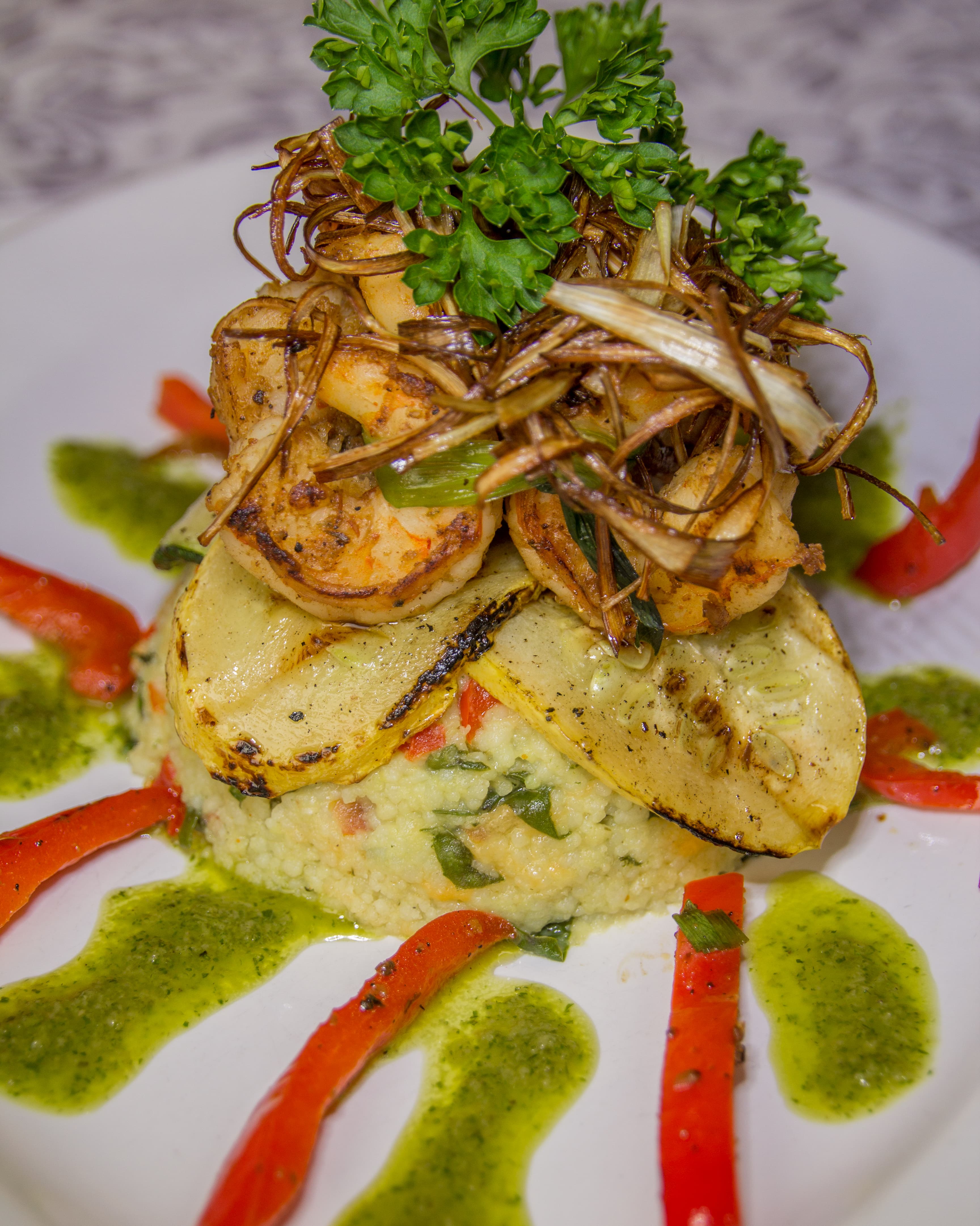 A gourmet dish featuring grilled shrimp on a bed of couscous, garnished with herbs and surrounded by red pepper strips and green sauce.