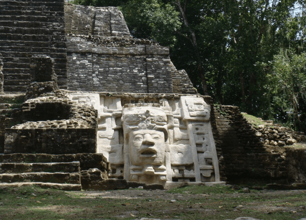 A stone carving of a face adorns a stepped pyramid in a lush, green archaeological site.