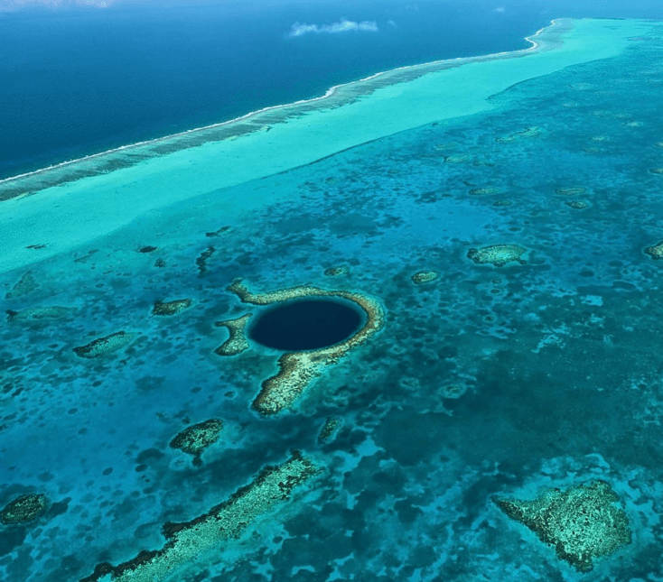 Aerial view of a deep blue circular sinkhole surrounded by vibrant turquoise waters and coral formations.