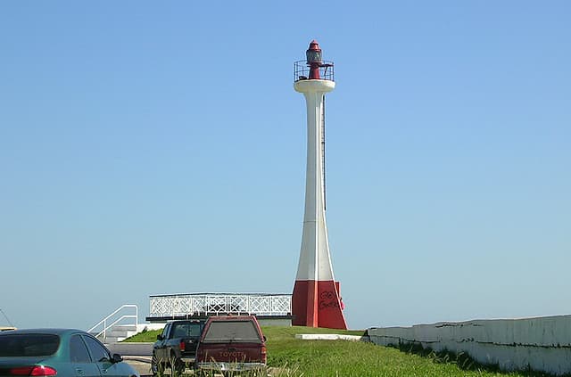 A tall white and red lighthouse stands beside a road with parked cars and a clear blue sky in the background.
